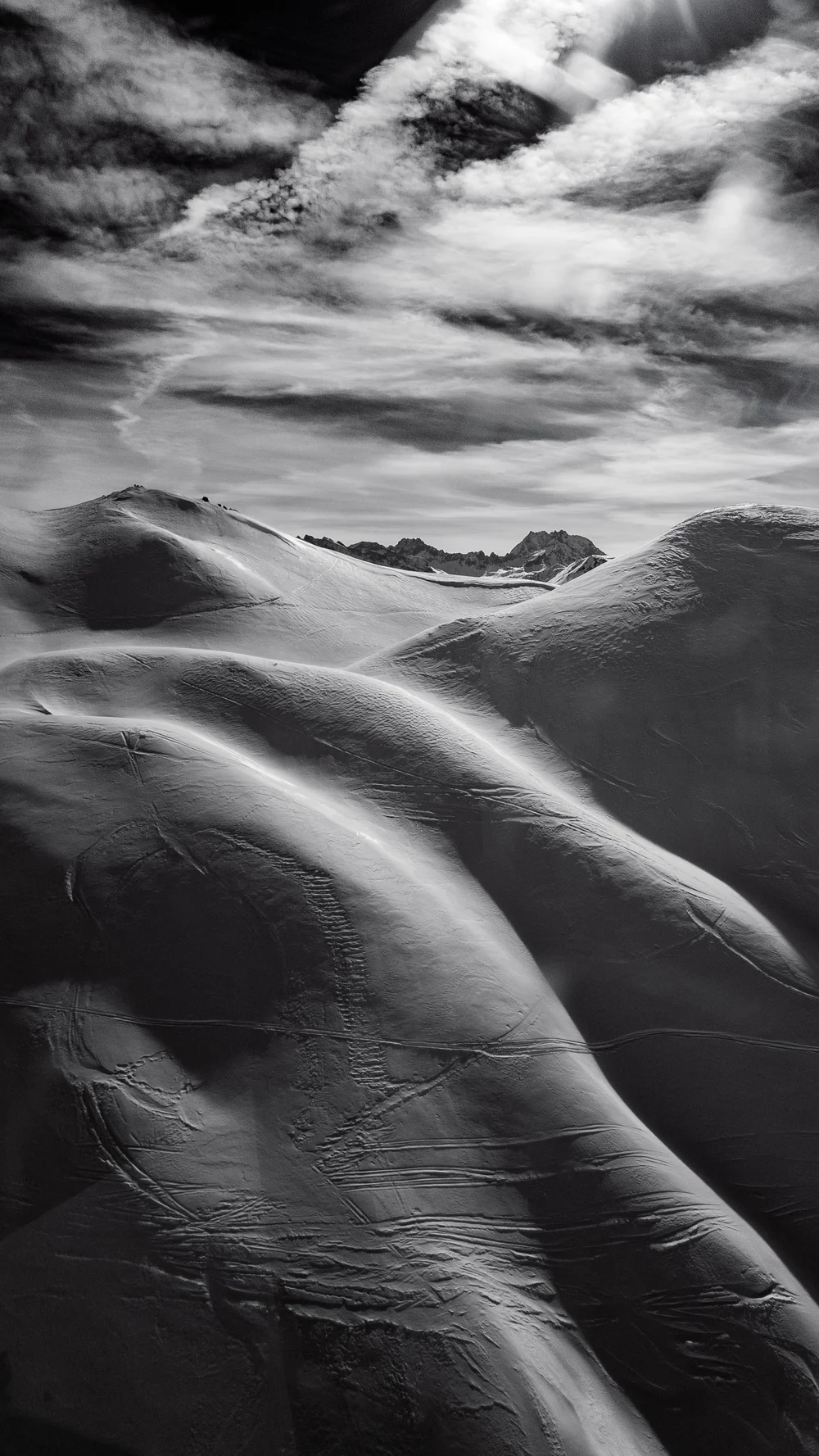 Black and white photo of smooth, curved snow-covered hills under a partly cloudy sky with distant jagged mountains.