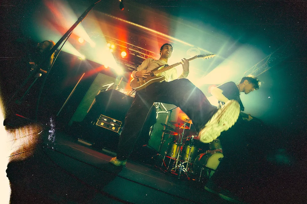 Two guitarists performing energetically on stage with bright colorful spotlights and drum set in the background.